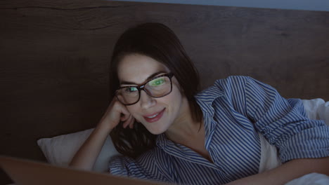 vista de cerca de una mujer sonriente usando una laptop y sonriendo mientras ve algo por la noche en su cama