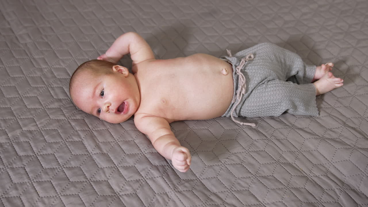 Lovely baby on the bed wearing pants only. Cute baby lying on the broad bed and sneezing. Infant kid on the grey backdrop view from above.