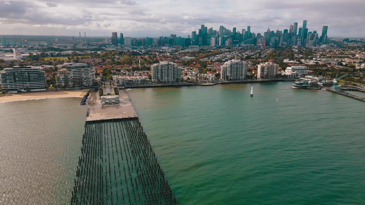 Riprese aeree panoramiche che mostrano il Princes Pier, case fronte mare, il CBD di Melbourne e un vasto paesaggio costiero durante una mattinata soleggiata e ventosa.