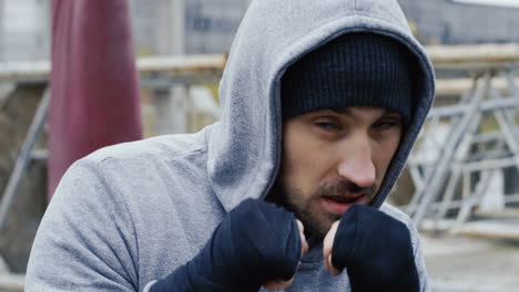 close-up view of caucasian bearded caucasian man in grey hoodie and boxing to the camera outdoors an abandoned factory on a cloudy morning