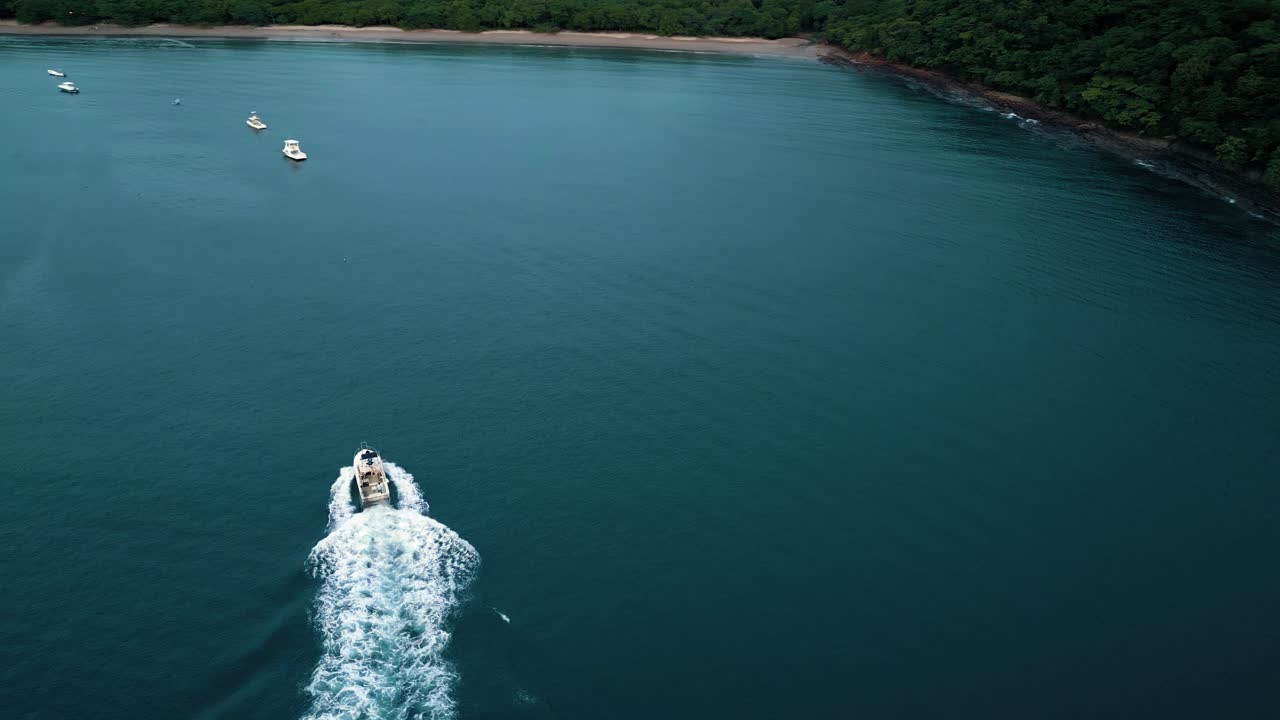 Ripresa aerea con drone di una barca a vela che naviga attraverso un golfo blu verso la riva, con altre barche sullo sfondo e vegetazione sulla costa