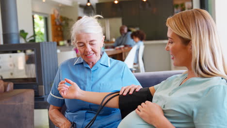 Senior-Midwife-Visiting-Pregnant-Woman-At-Home-Taking-Blood-Pressure-With-Family-In-Background