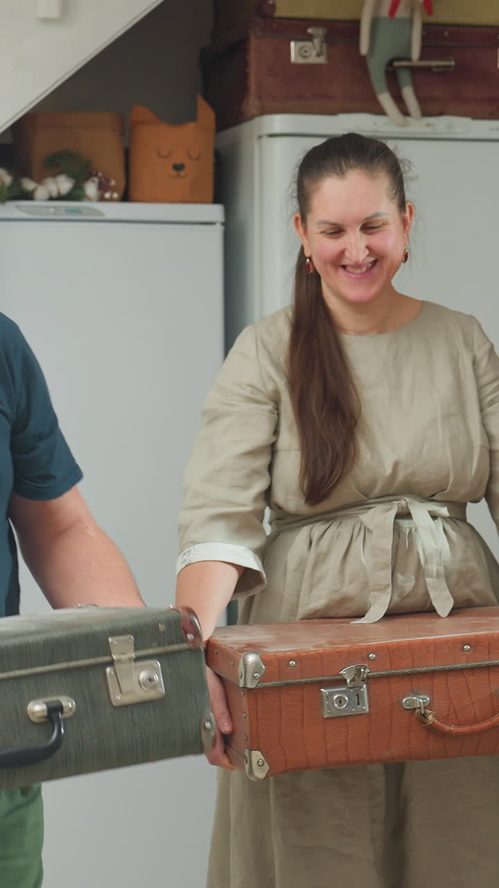 Casal caucasiano sorrindo com malas na cozinha: mulher sorridente vestindo um vestido de linho segura uma mala marrom, enquanto um homem sorridente segura um baú verde; interior iluminado com geladeira e prateleiras ao fundo