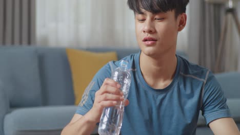 스포츠 옷을 입은 아시아 남성이 아침 운동을 마친 후 집에서 미니멀한 인테리어를 가진 넓고 밝은 아파트에서 물을 마시고 있습니다.
