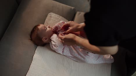 Top-view-of-a-little-girl-baby-lying-on-a-special-mat-on-a-gray-sofa-while-her-mother-fastens-the-buttons-on-the-little-girls-overalls