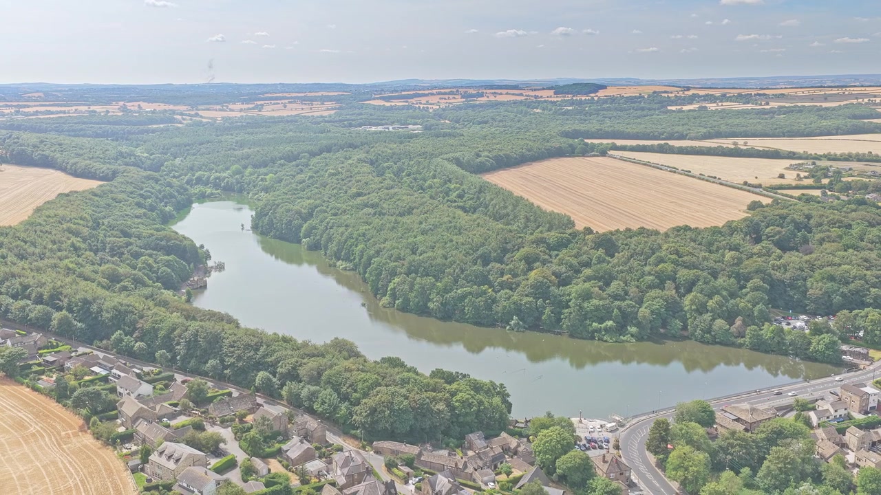 El embalse y el bosque de Newmillerdam bordean la carretera A61 Barnsley cerca de Wakefield, West Yorkshire, Inglaterra, con tierras de cultivo y viviendas al fondo, toma de dron en retroceso