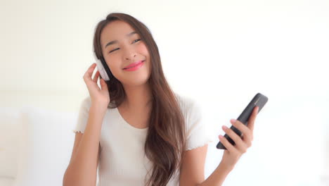 Portrait-shot--Asian-girl-listen-to-music-from-her-overhead-Bluetooth-headphones-and-holding-a-mobile-phone-in-one-hand-dancing