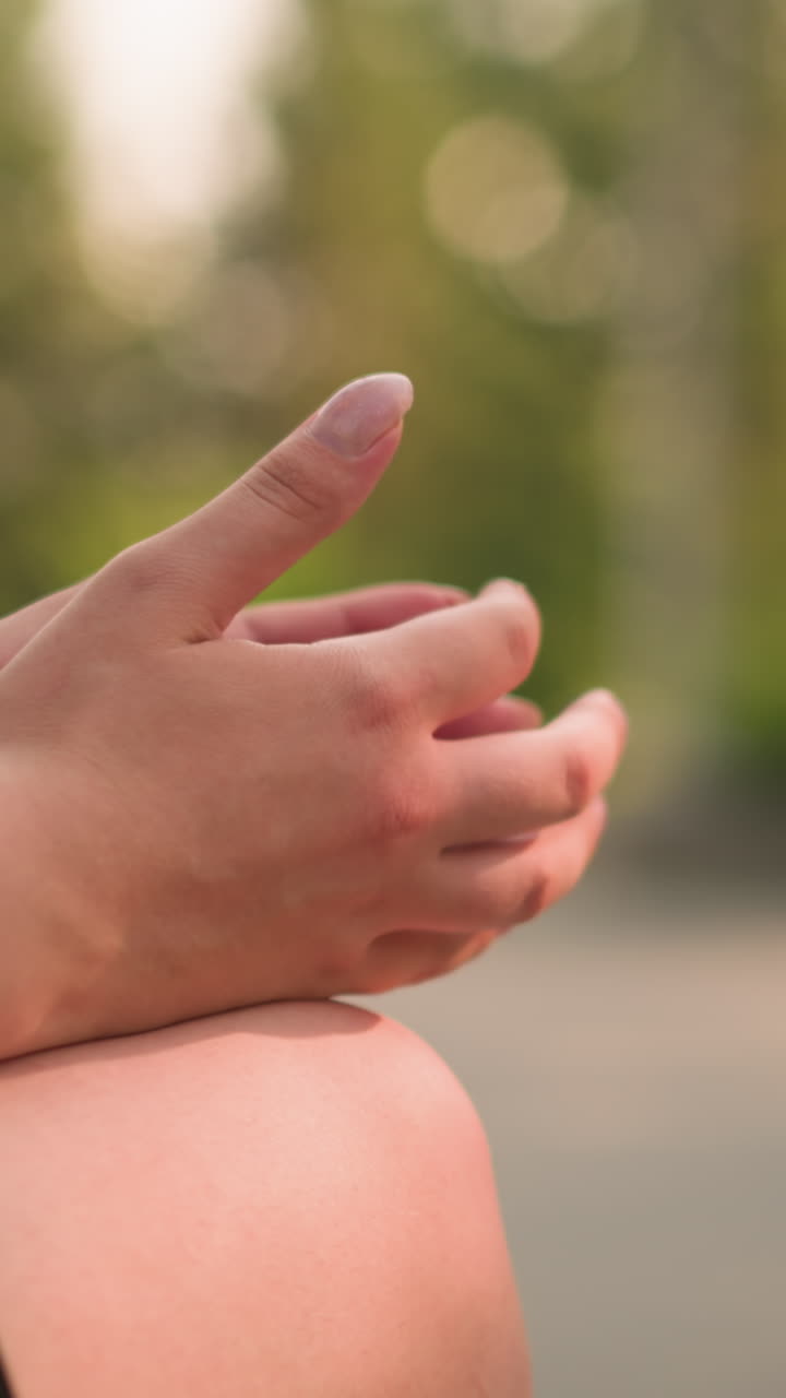 vista parcial de individuos sentados al aire libre con las piernas cruzadas, la mano apoyada en la rodilla apretando suavemente la otra mano bajo la cálida luz solar, fondo suave y borroso con vegetación