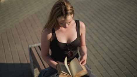 dama sentada al aire libre con una camiseta negra con gafas de sol colgando de ella, hojeando páginas de un libro mientras está enfocada, el fondo presenta pavimento estampado, y parece profundamente pensativa