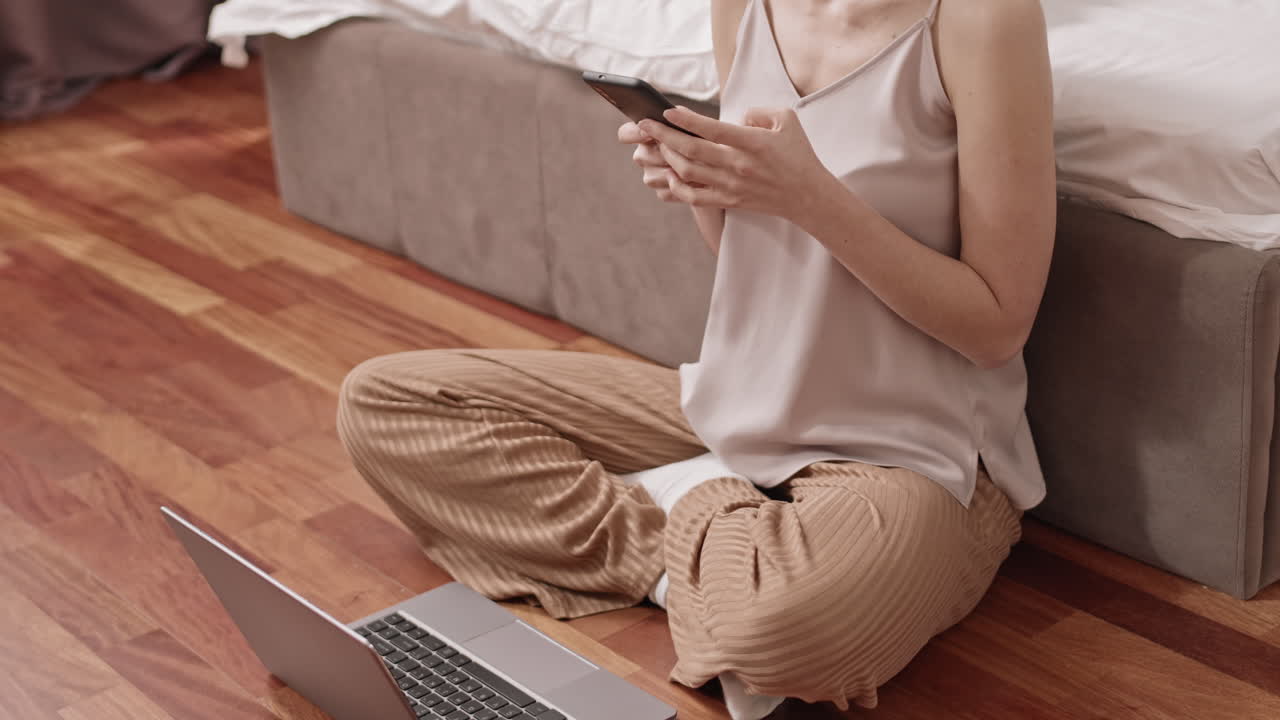 Woman sitting on floor using phone and laptop in bedroom