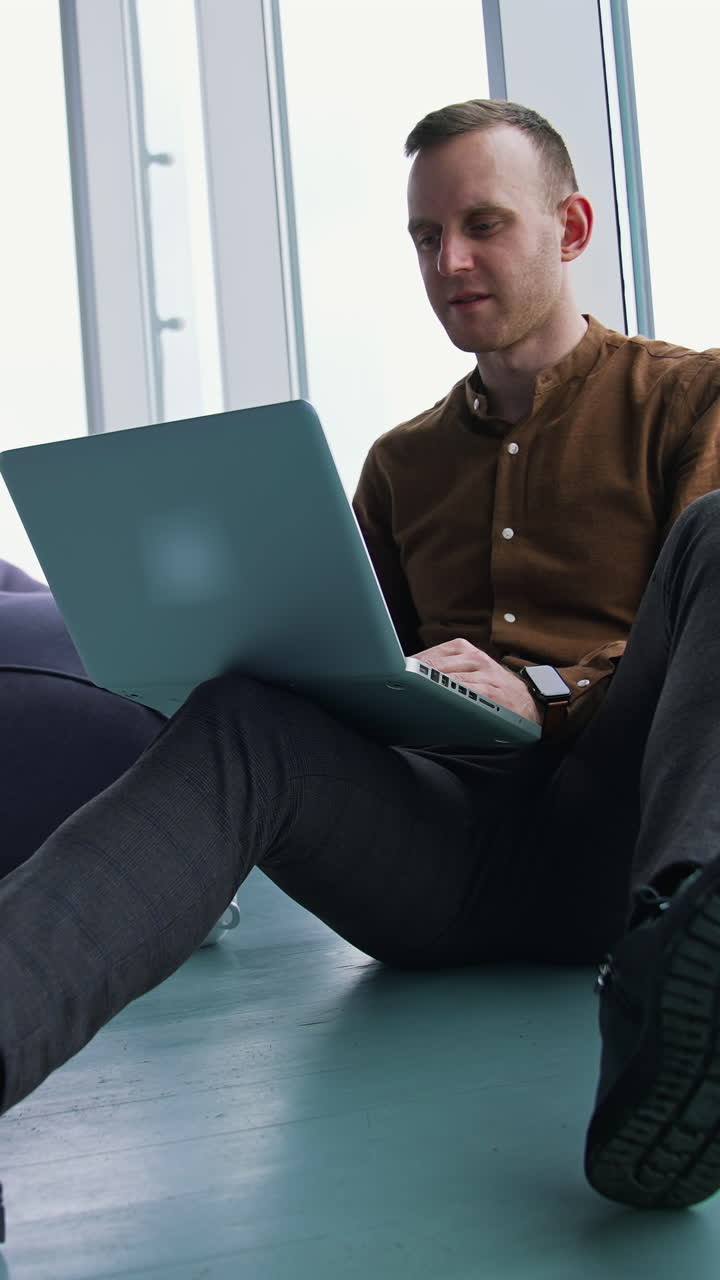 Relaxed businessman in modern office. Young man is sitting on a floor and working on a laptop on windows background. Balanced work. Vertical video