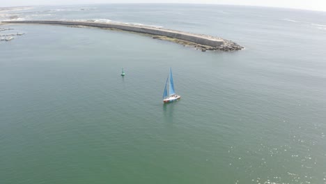 Drone-orbits-sailboat,-revealing-coastal-cityscape-on-a-sunny-day-with-fish-in-the-water