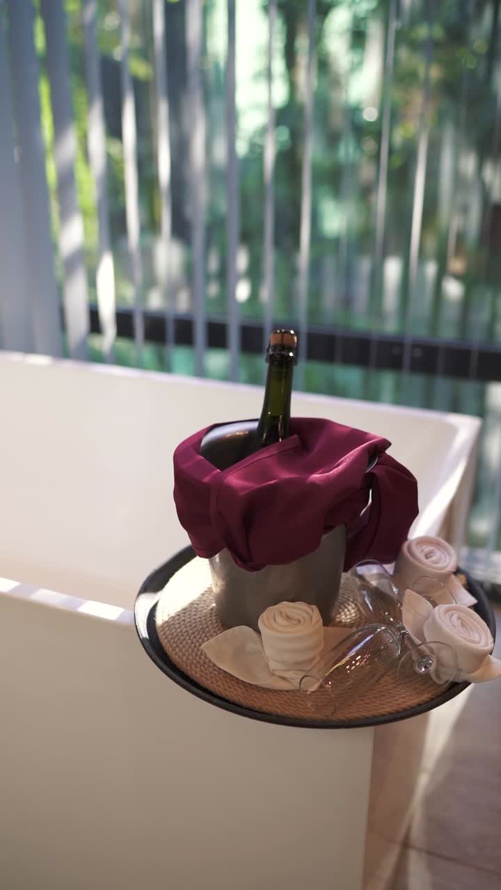 Slow moving vertical shot of champagne bottle and glasses on tray in bath room at hotel, with large window in back and white bath tub.
