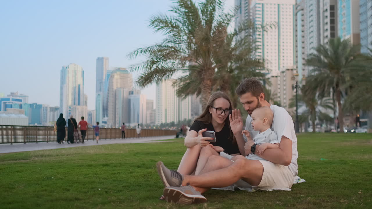 Happy family with two children sitting together on grass in park and taking a selfie. With smartphone.