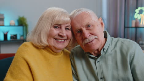 Happy-old-senior-elderly-family-couple-hugging,-laughing,-smiling-looking-at-camera-at-home-sofa