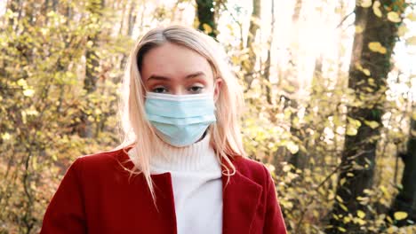 portrait of a young beautiful woman putting on corona safety mask amidst orange brown autumn forest woodland while wearing a red coat version 3