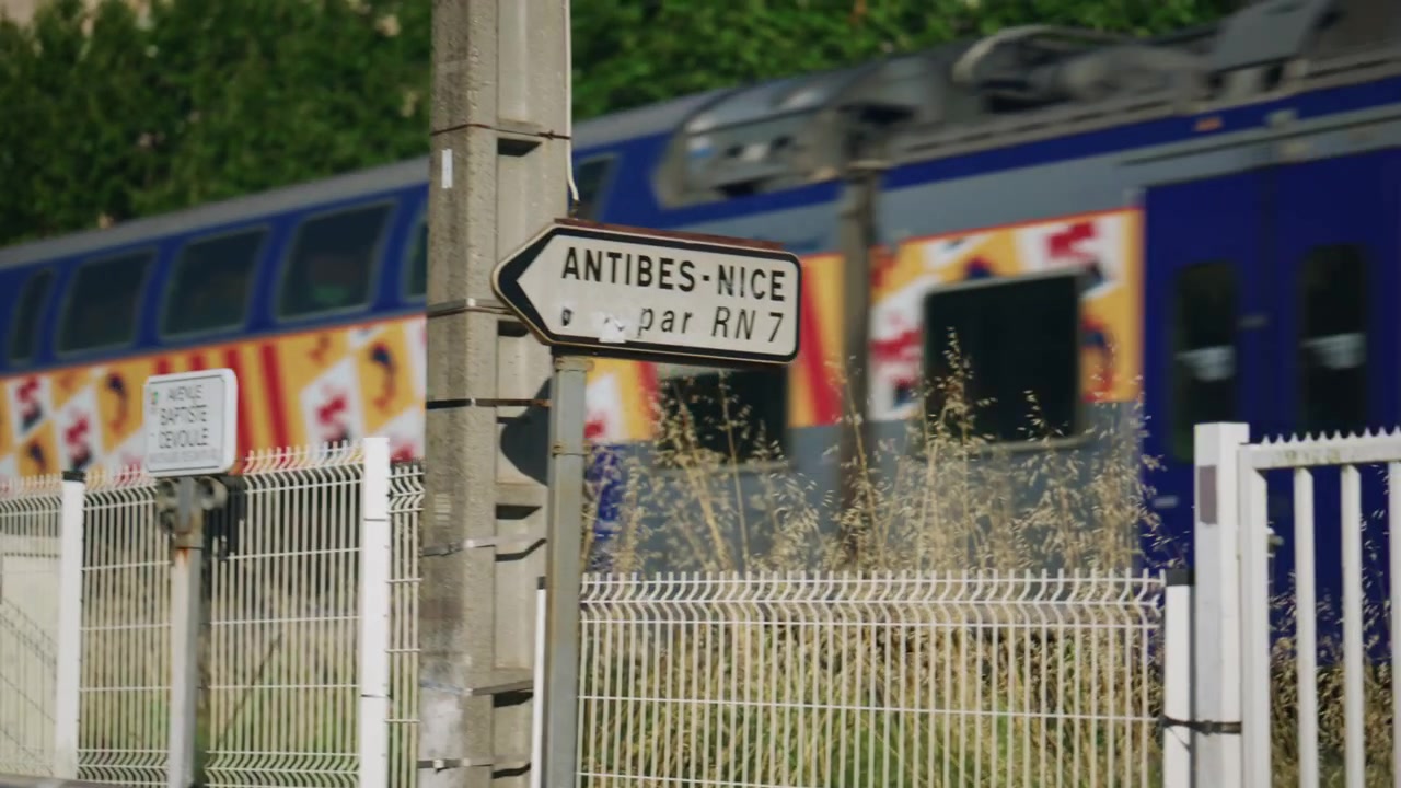 Tren en movimiento pasando detrás de una barandilla azul, con gráficos exteriores coloridos y vegetación en primer plano, creando una dinámica escena de viaje urbano, Golfe Juan Vallauris, Francia.