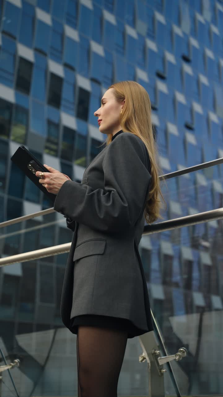 Real estate agent caucasian businesswoman in a gray blazer stands confidently outdoors, working using tablet near a modern glass office building