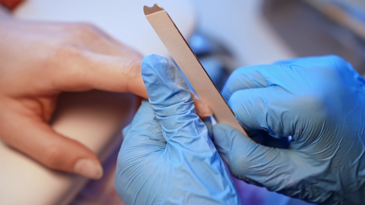 Close-up of a gloved nail technician shaping a client’s natural fingernail with an emery board during precise professional manicure preparation.