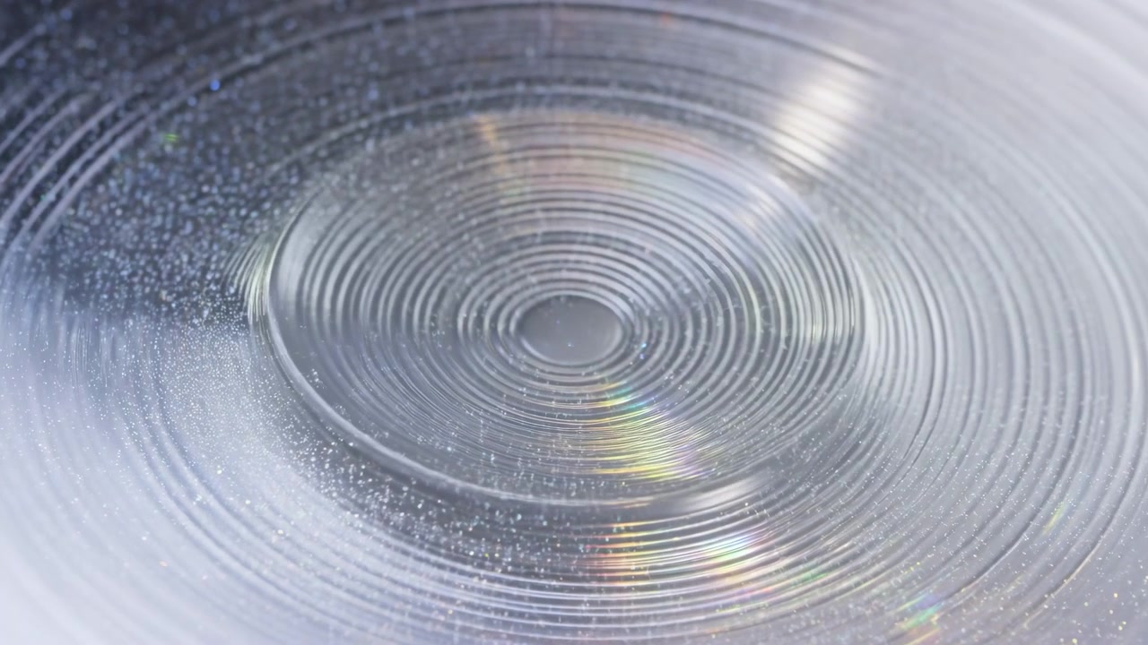 Una vista macro segue scanalature circolari fitte attorno a un disco centrale, mentre luce argentea, piccoli dettagli maculati e riflessi prismatici si muovono su una texture liscia e traslucida