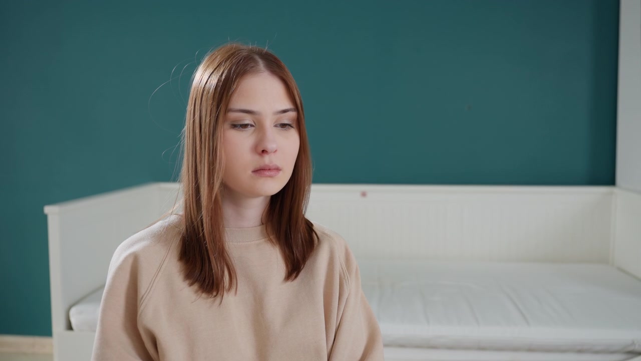 Young white woman sitting on bed listening to online grammar tutor, pensive expression, cozy bedroom with teal wall, oversized sweater, soft natural light, quiet study mood, subtle concentration