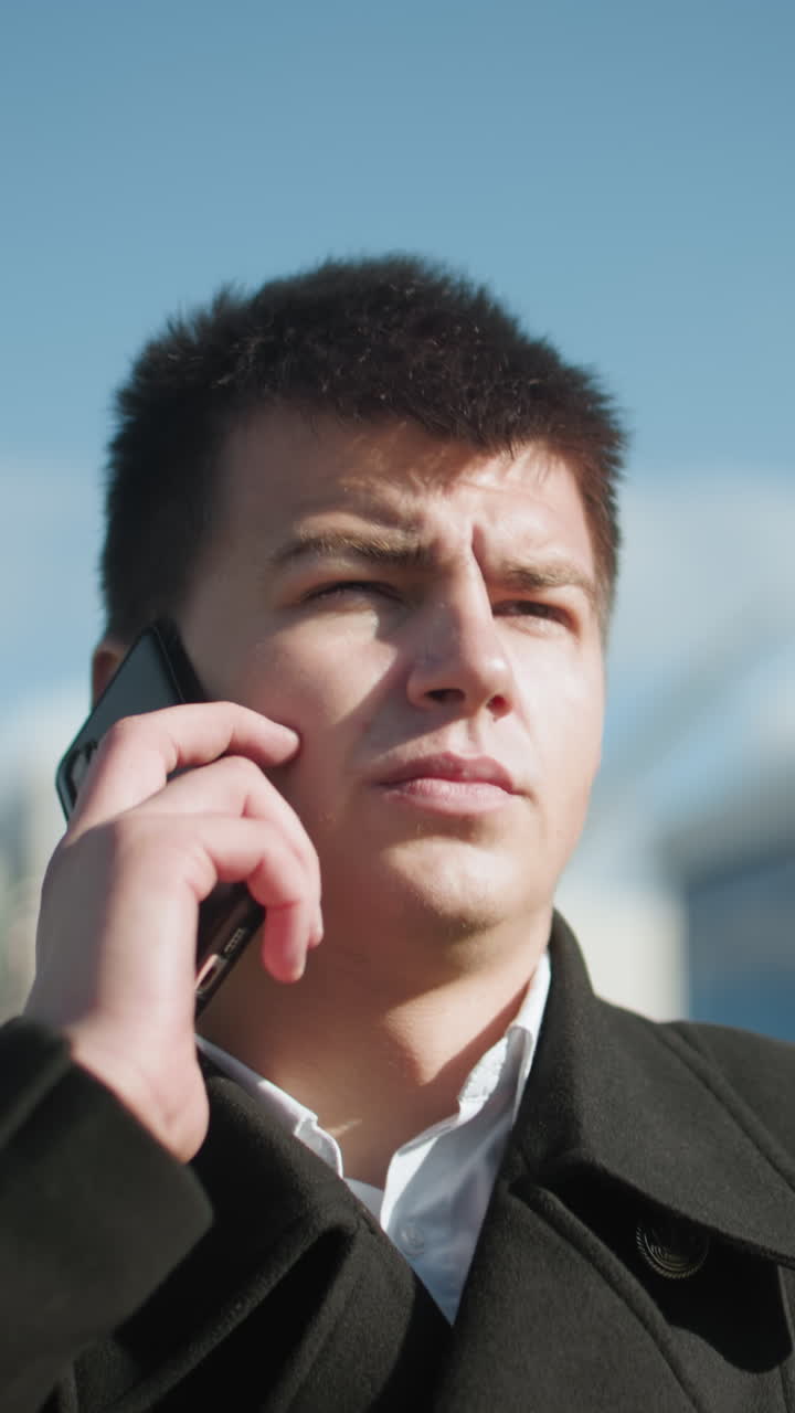 검은 코트를 입은 투자자가 도시 환경에서 야외에서 전화 통화에 응답합니다. 배경은 고 밝은 빛 아래 흐릿한 현대 건물과 초록색으로 전문성을 발휘합니다.