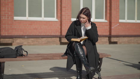 young lady seated on a bench reading her book while adjusting her hair, with one leg crossed, two bags are placed on the bench, and buildings are visible in the background