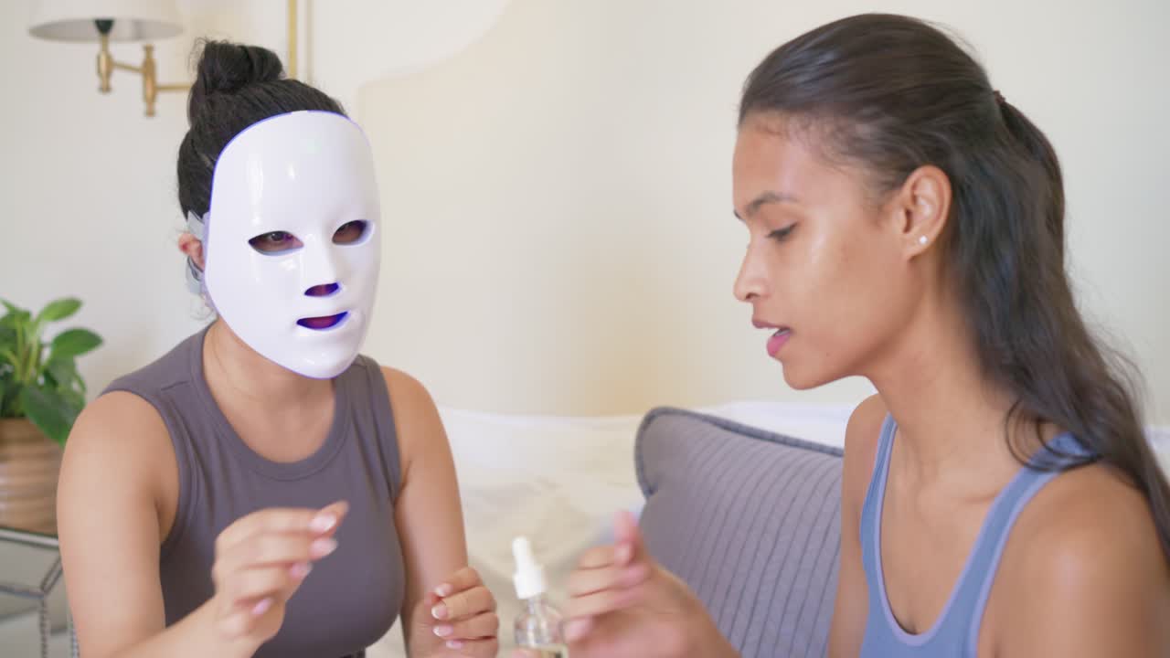 Amiche di varie etnie applicano una maschera facciale a LED, condividendo un contagocce di siero durante la routine di cura della pelle sul letto.