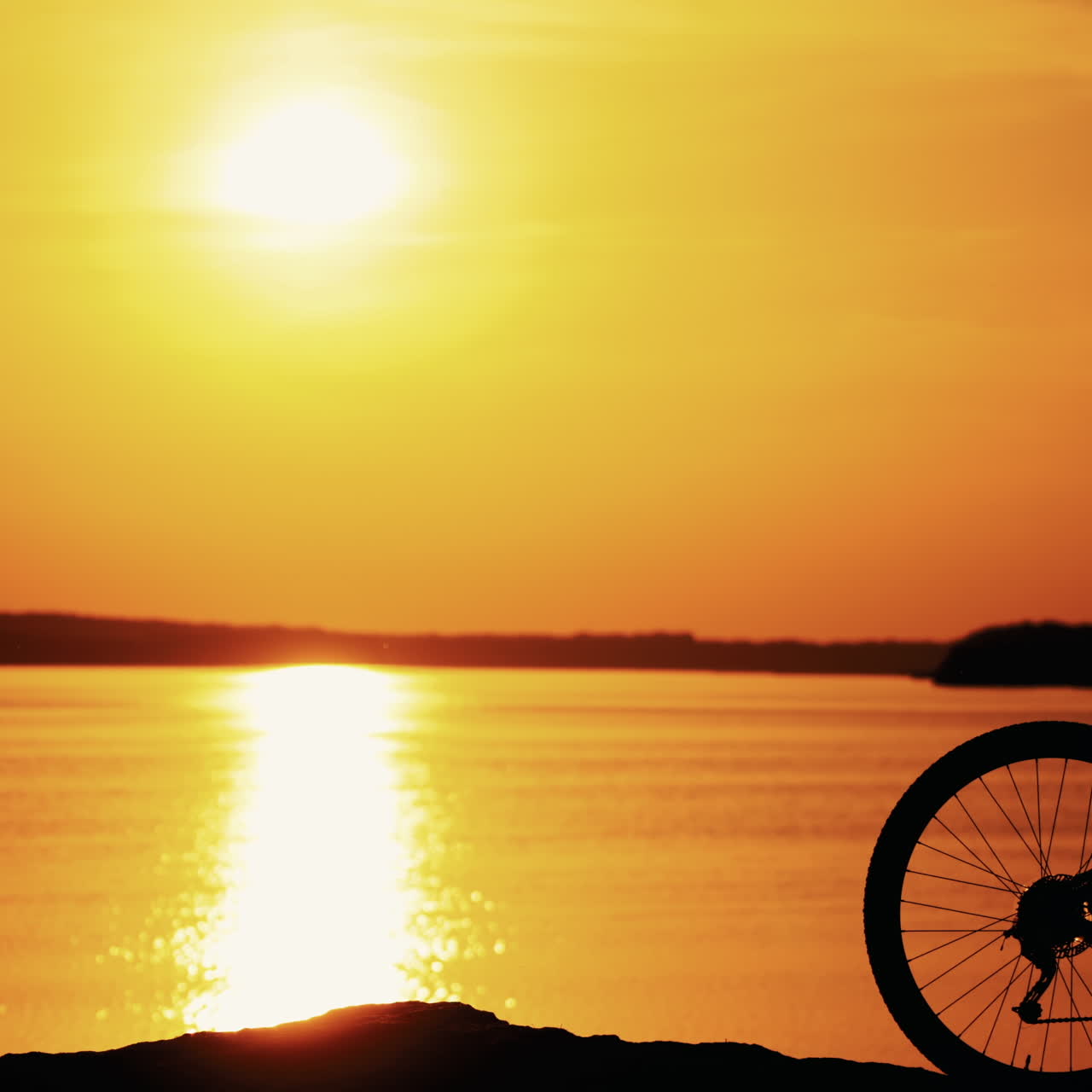 Silhouette of a professional cyclist with helmet and glasses at sunset. Bike rider is putting on his protective glasses and moves further on the background of the golden path from the sun over the water surface.