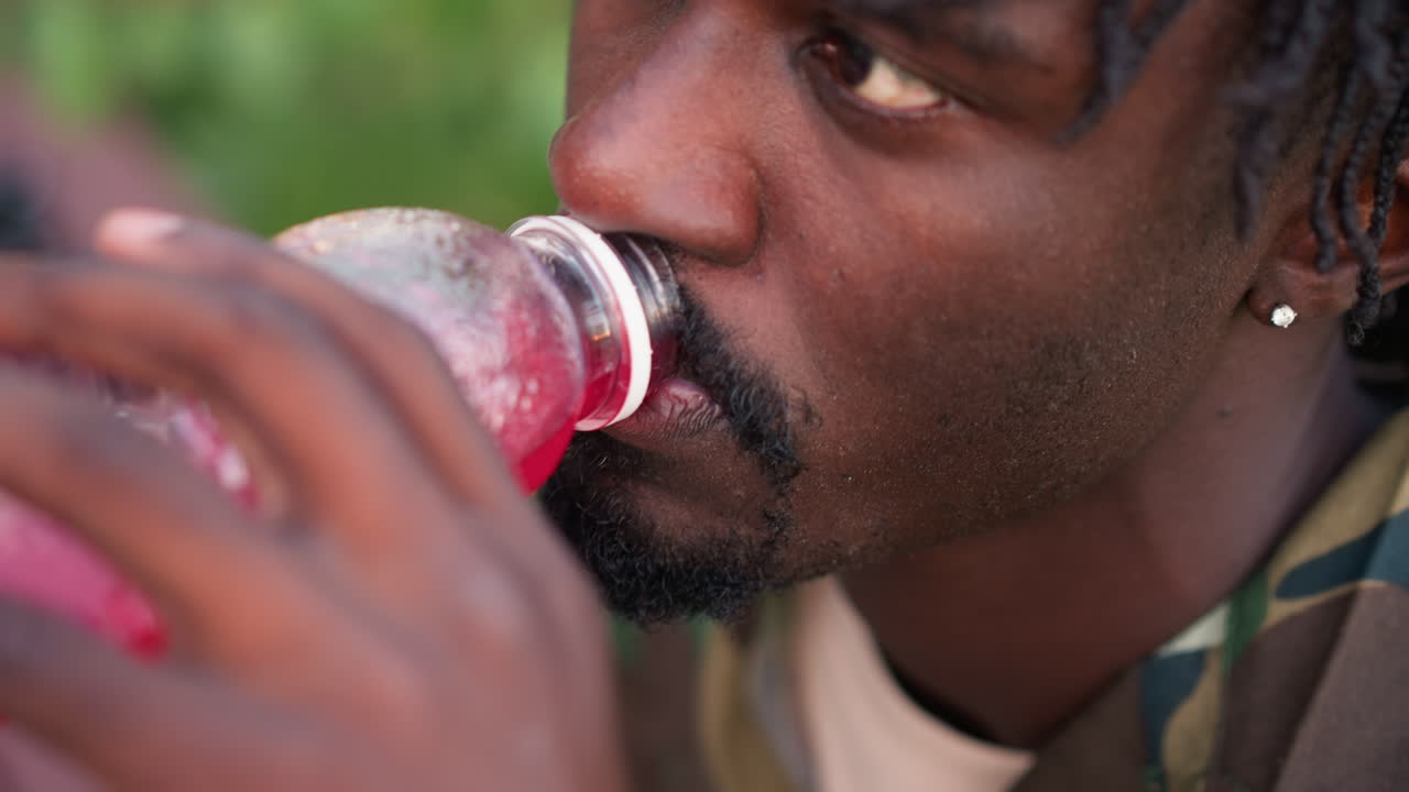 Camouflage Jacket Man Sipping Red Drink Outdoors, Closeup Profile Showing Focused Gaze, Sweaty Skin, Stubble, Hand Gripping Bottle, Subtle Earring, Relaxed Patrol Break Atmosphere, Soft Natural Light