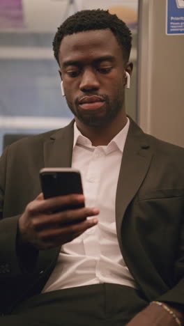 vertical video of young businessman in suit commuting to work sitting in london underground tube train wearing wireless earbuds to stream from mobile phone shot in real time 3