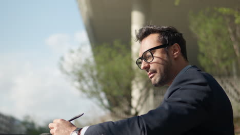 Profile-of-a-man-in-suit-working-outdoors-in-a-park-eating-snacks-and-writing-with-pen---low-angle-panning