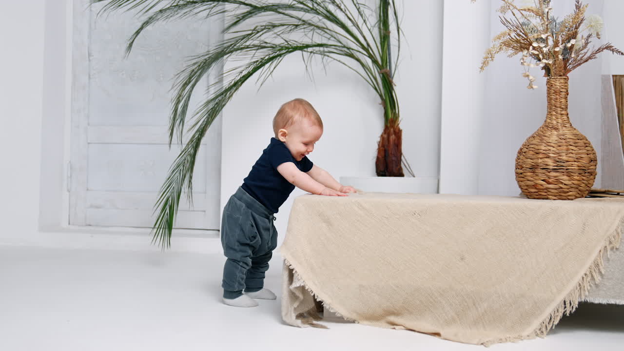 Lovely cute baby boy stands by the coffee table tapping by the surface. Infant kid trying to walk himself.