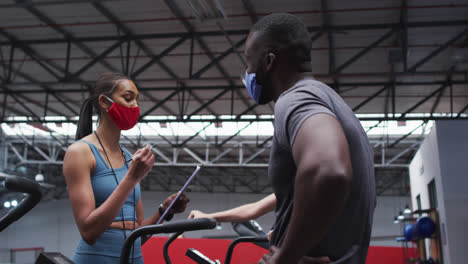 Mixed-race-female-fitness-trainer-wearing-face-mask-at-gym