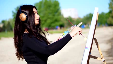 summer, near river, on beach. at sunrise, a beautiful woman artist holds a brush in her hand, draws a picture. she paints on canvas god shiva. listening to music on headphones