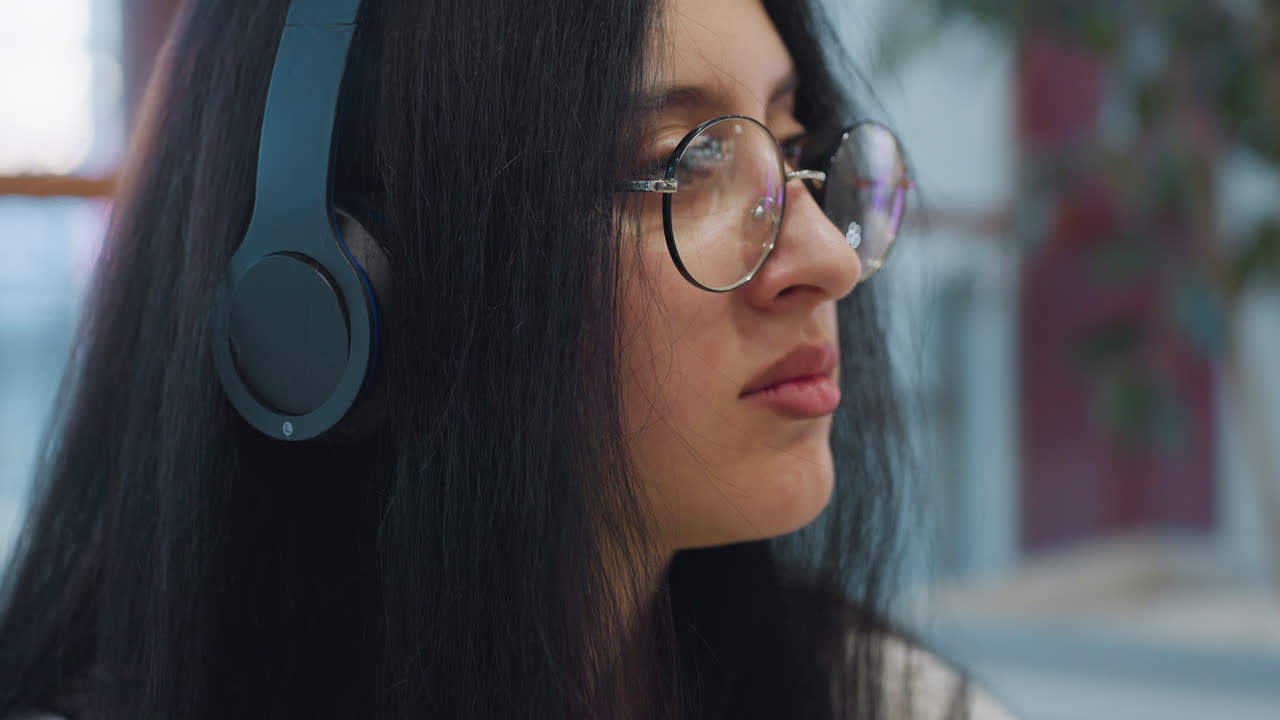 Close up of young woman with long dark hair wearing large headphones and glasses gazing thoughtfully to side in soft indoor lighting, portraying calm moment of reflection or deep thought