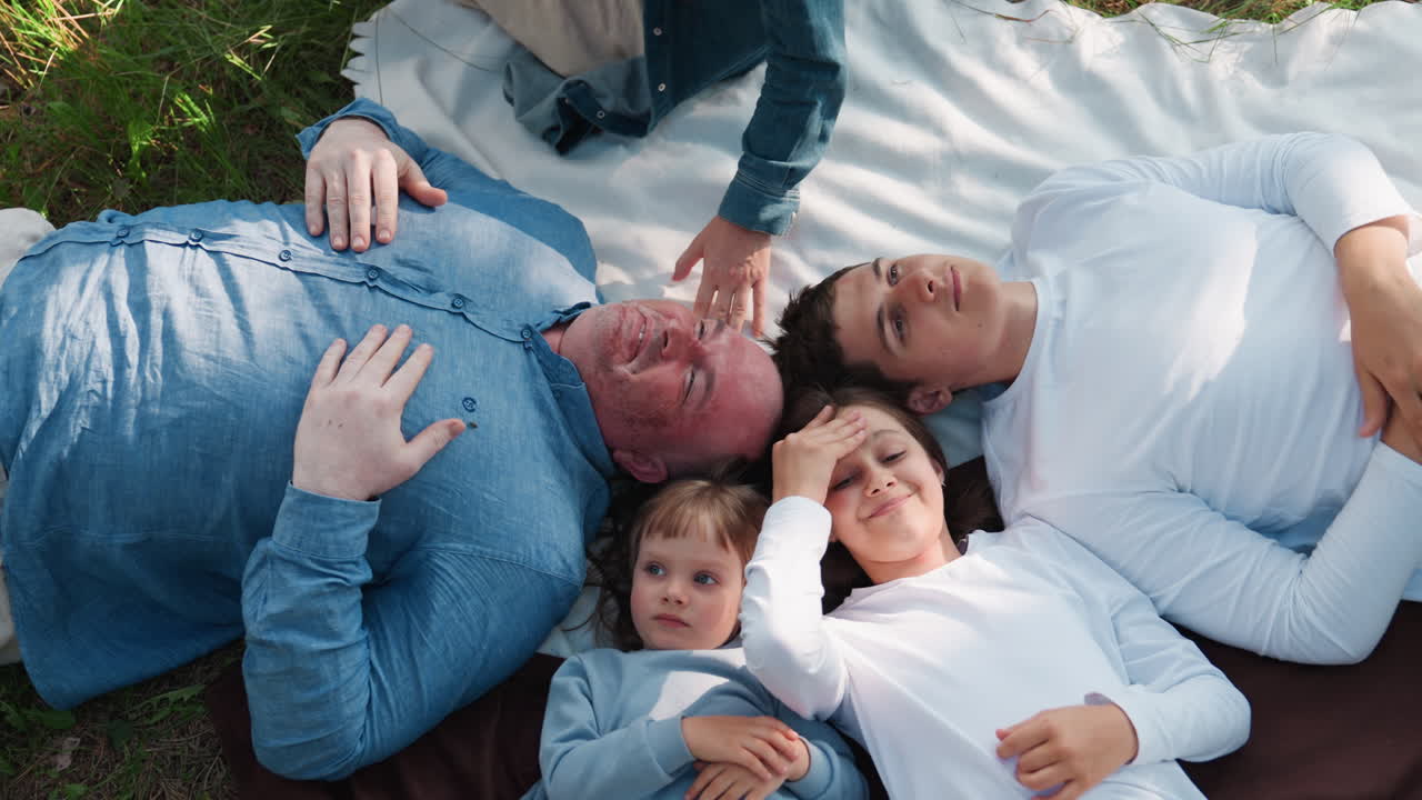 Happy family lying faced up during picnic on soft blanket outdoors, enjoying peaceful relaxation together in sunlight, expressing love, unity, warmth, and calm connection between parents