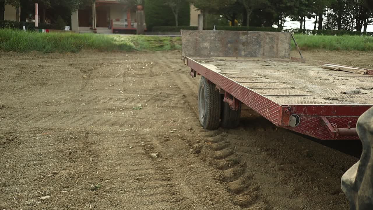 Close up footage of big wheels of green agricultural tractor and red trailer on a prepared soil