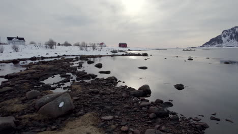 Flying-over-froozen-ocean,-rocks-and-water-with-the-sun-reflecting-on-the-surface,-revealing-a-Norwegian-Farm