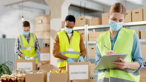 front view of caucasian female volunteer in facial mask checking donation list and smiling to the camera in warehouse house