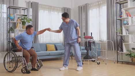 full body side view of asian male nurse holding patient's hand who in a wheelchair to stand up and walk during a physical therapy at home