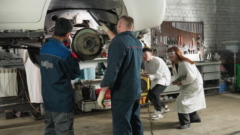 engineer working on car repair while lab technicians approach with tablet to observe and take notes, mechanical workshop setting with tools, lifted vehicle, teamwork, technical inspection