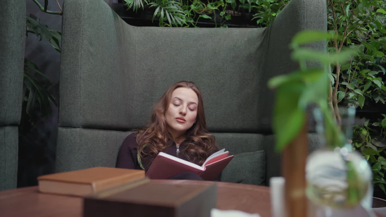 Blonde woman wearing black top sits in green cushioned booth writing thoughtfully in pink notebook, with indoor plants and soft lighting creating calm atmosphere, tabletop elements visible