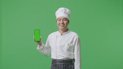 el chef asiático sonriendo y mostrando el teléfono inteligente de pantalla verde a la cámara mientras está de pie en el estudio de fondo de pantalla verde