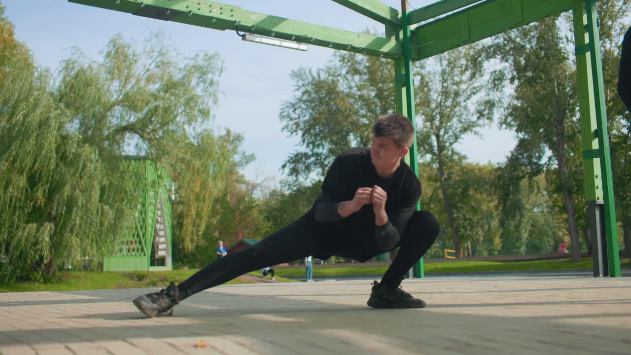 young man stretching under green pavilion, performing deep lateral lunges and controlled holds while companion stands on scooter, morning sunlight, leafy trees, focused expression, black sportswear