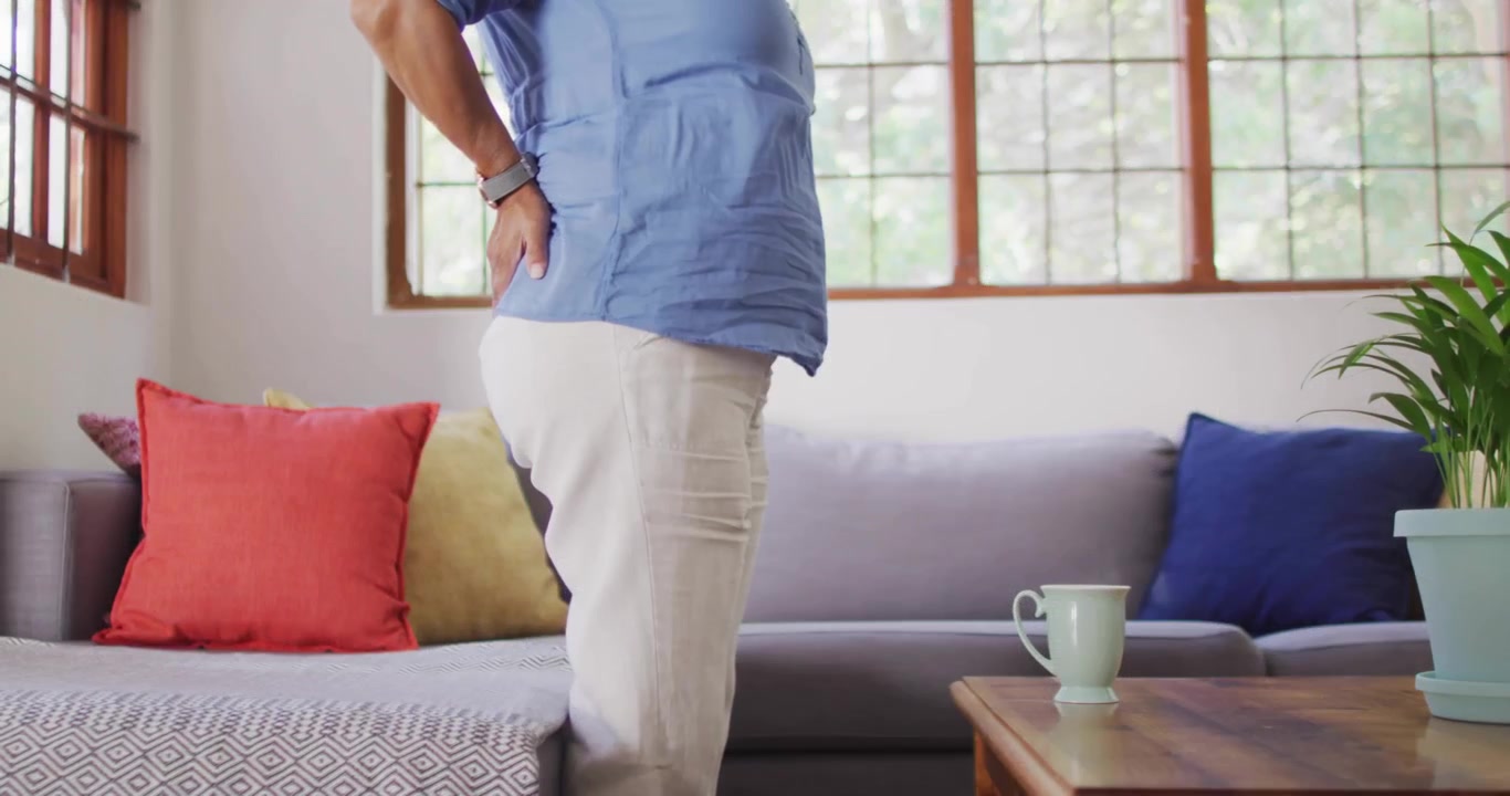 Senior woman experiencing back pain gripping ottoman and sofa while performing posture correction