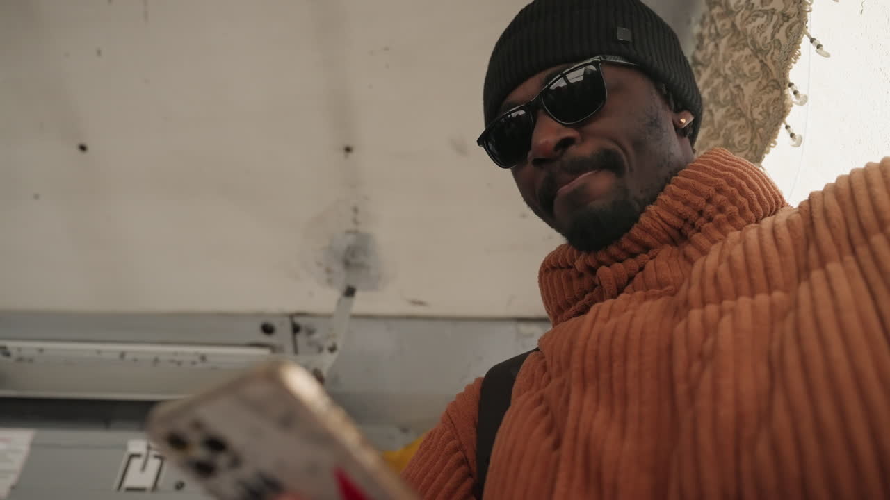 Ritratto in transito da angolazione bassa di un uomo che controlla il telefono mentre sale sull'autobus, cappotto di velluto a coste arancione, berretto e occhiali da sole creano un audace look streetwear, movimento spontaneo e luce diurna ambientale, soffitto metallico e corrimano.