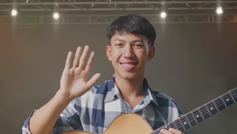 primer plano de un adolescente asiático con la mano agitando la guitarra y sonriendo a la cámara en el escenario
