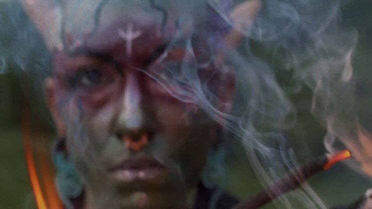 Person with Beltane ritual facepaint holds torch of fire and smoke near lens