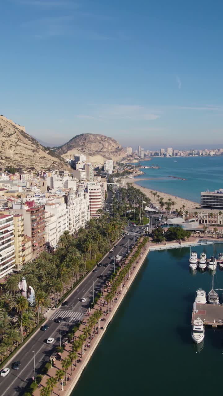 Filmato aereo verticale realizzato con drone che mostra il lungomare lungo il porto turistico di Alicante, con barche ormeggiate e lo skyline della città sul mare, Spagna